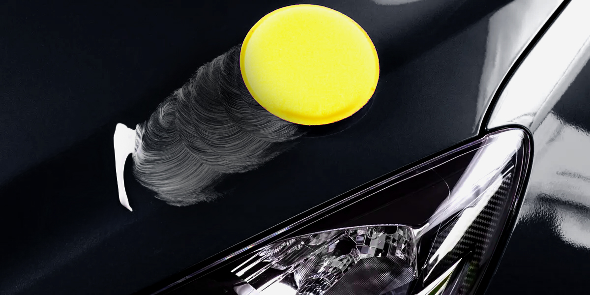 Applicateur en mousse jaune appliquant de la cire sur la surface du capot d’une voiture noire, près du phare avant, avec des mouvements circulaires visibles dans la cire.