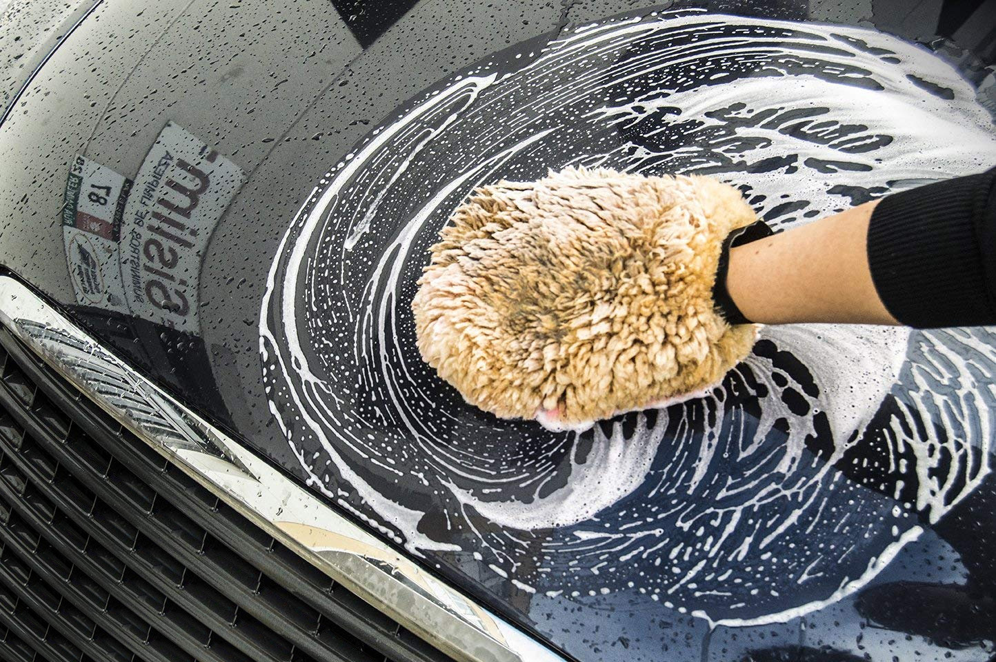 Main d’une personne utilisant un gant en laine d’agneau pour laver la peinture d’une voiture foncée couverte de mousse de savon