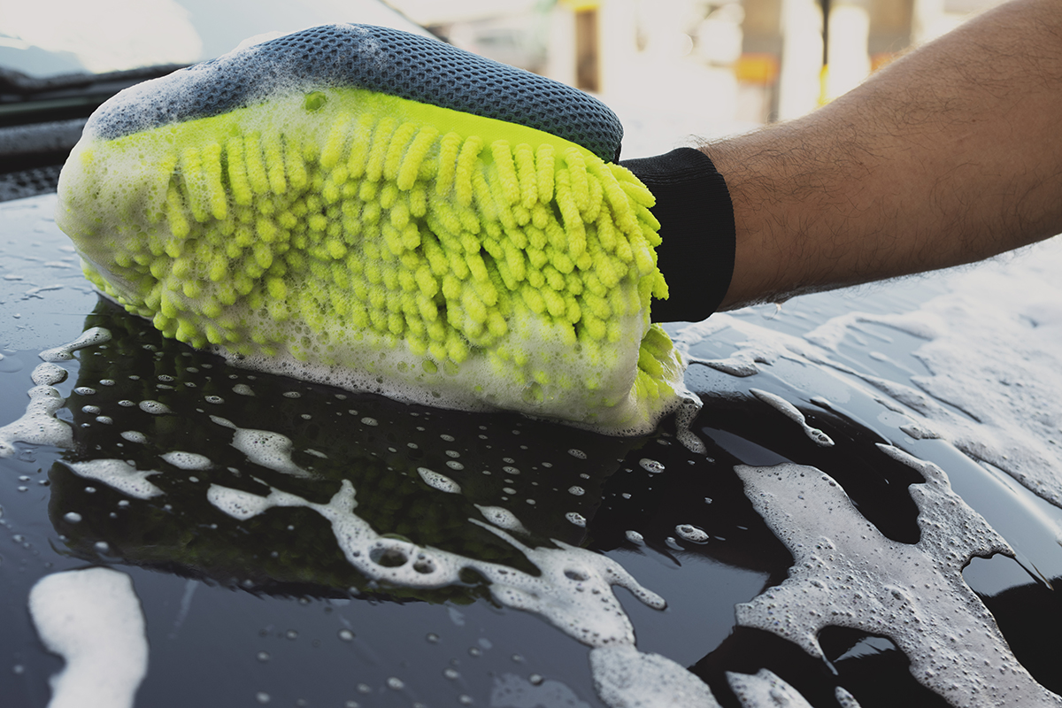 Une personne utilisant un gant en microfibre jaune rempli de mousse pour laver la surface d’une voiture noire, couverte de savon et d’eau.