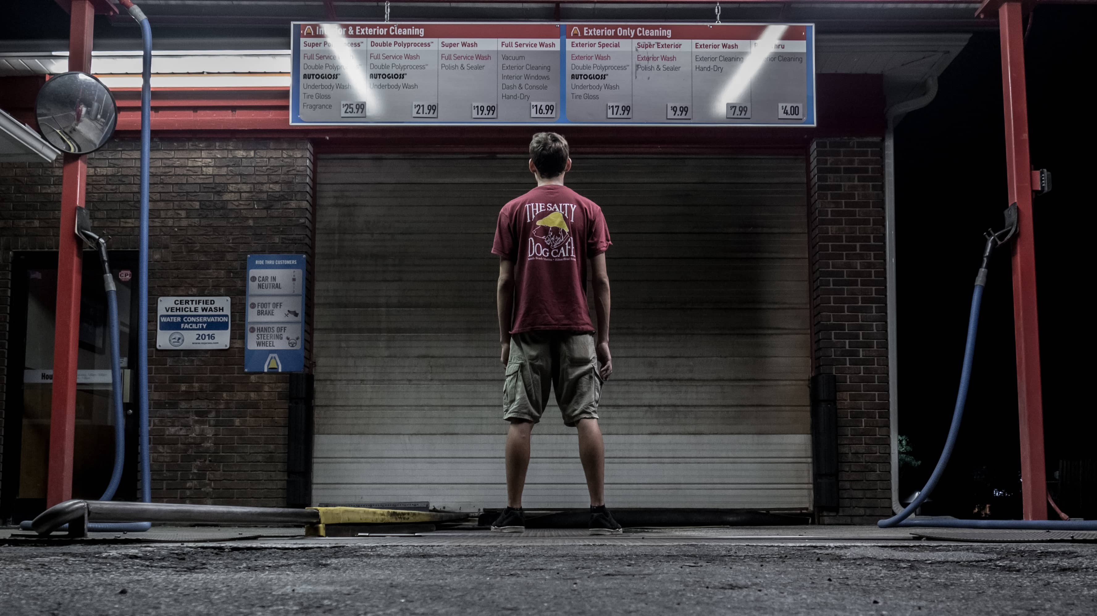 Personne debout dans une station de lavage automobile, observant un panneau de tarifs des services de nettoyage extérieur et intérieur. La personne porte un t-shirt rouge avec le logo « The Salty Dog Cafe » et un short cargo beige.