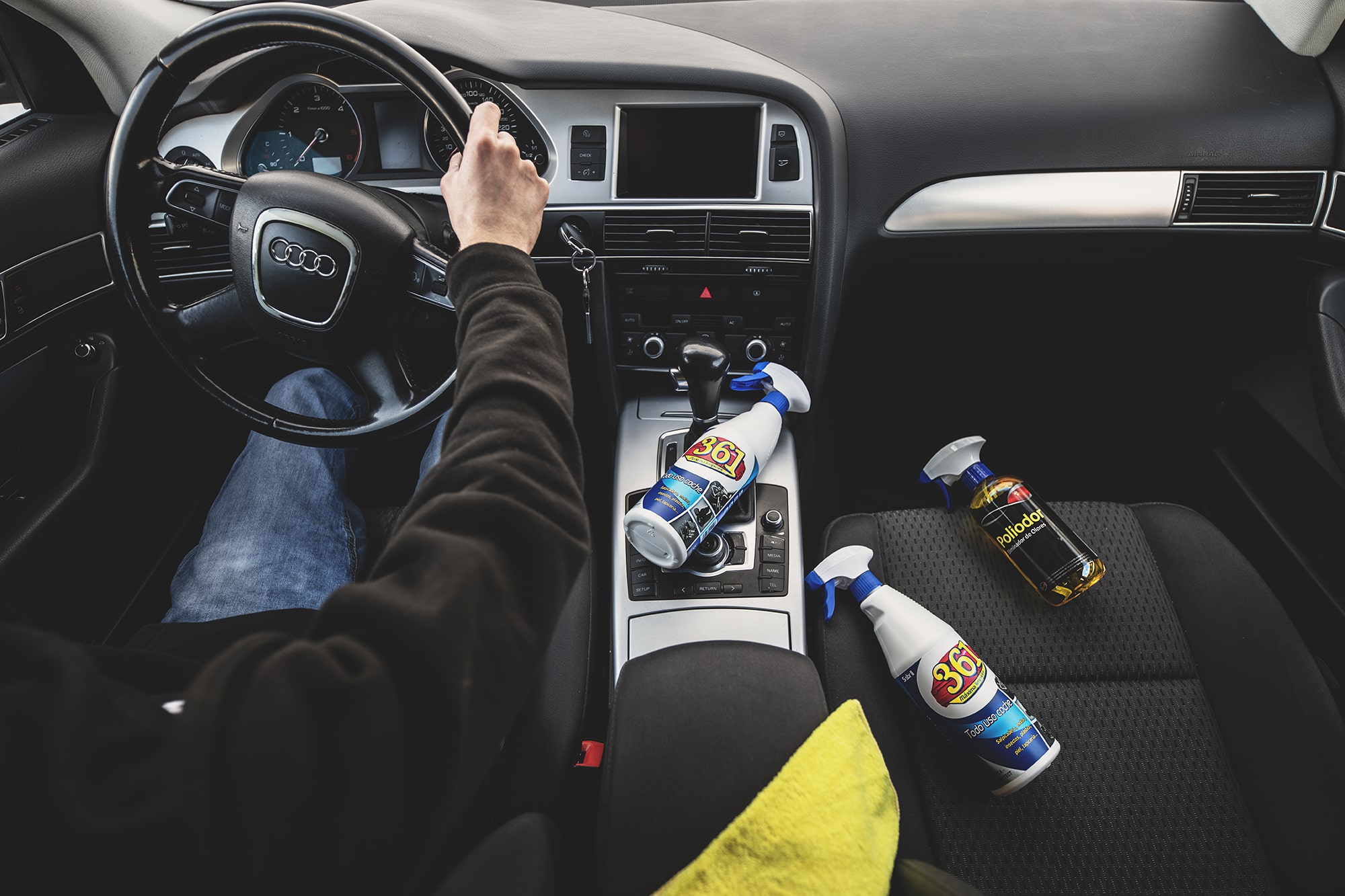 Intérieur d’une voiture Audi avec un homme au volant, tandis que sur le siège passager se trouvent deux flacons de 361 Todo Uso Coche et un Poliodor Éliminateur d’Odeurs.