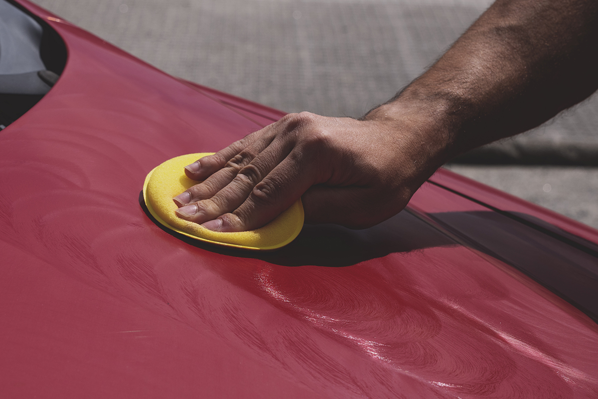Main d’un homme avec un applicateur en mousse jaune sur le capot d’une voiture rouge