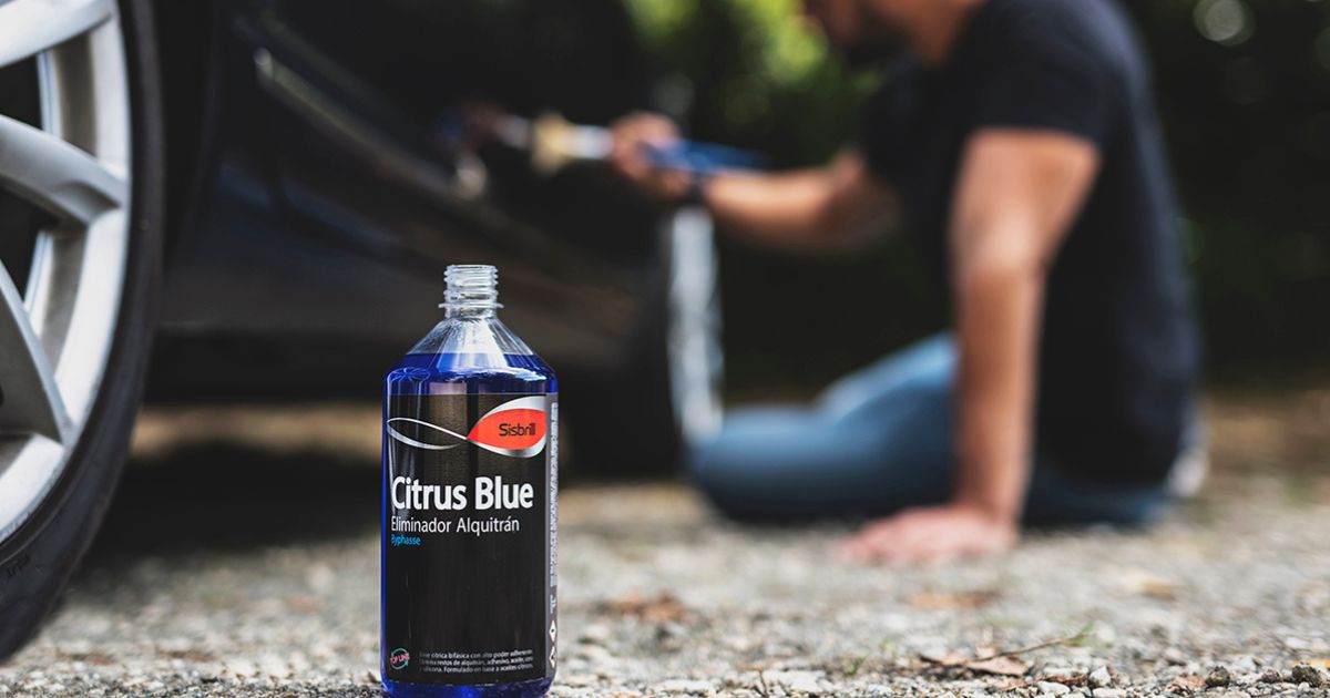 Une bouteille d’éliminateur de goudron Sisbrill Citrus Blue est au premier plan, posée au sol près de la roue d’une voiture, tandis qu’en arrière-plan, légèrement floue, une personne semble appliquer le produit sur le véhicule à l’aide d’un pinceau.