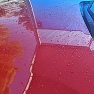 Capó de coche rojo con gotas de agua que muestran un efecto de repelencia, posiblemente tras la aplicación de cera protectora, reflejando árboles y un edificio cercano.